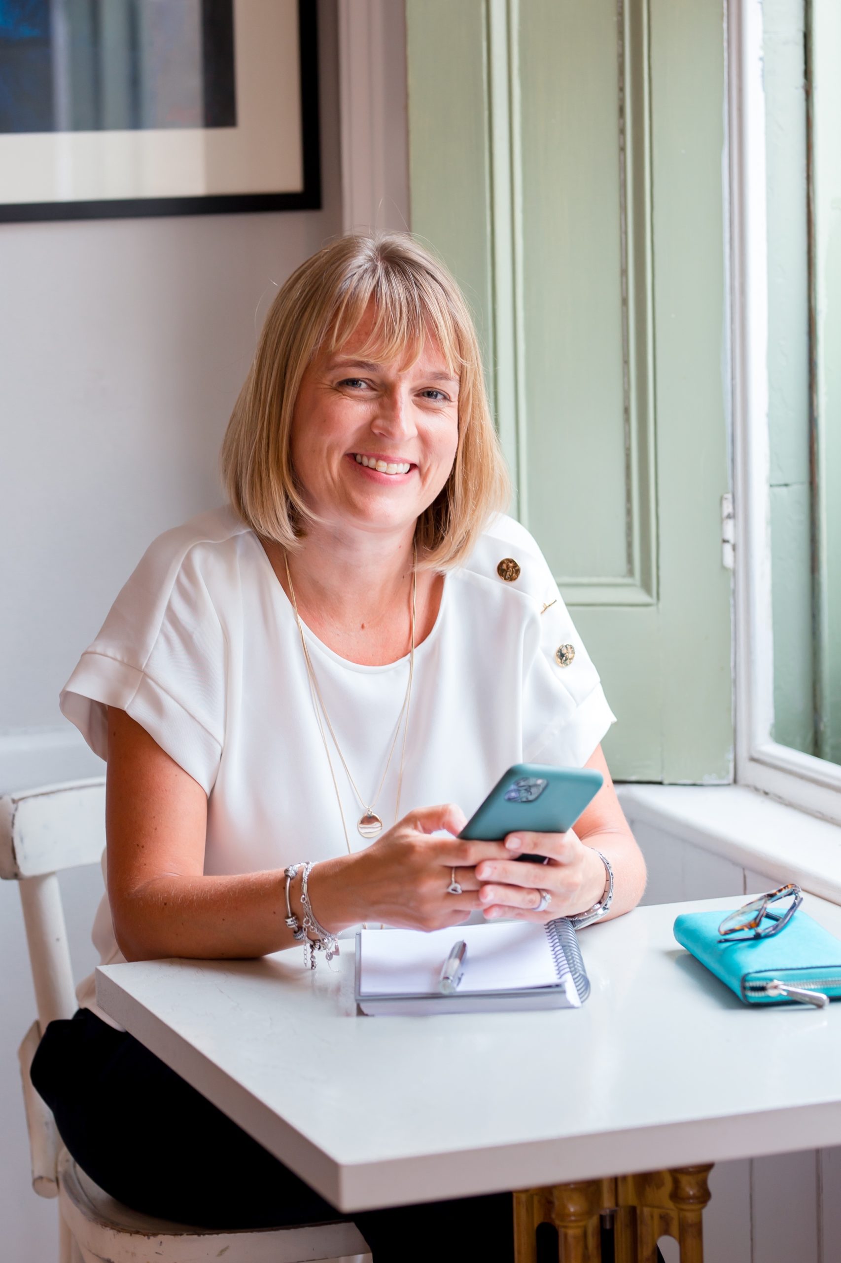 Picture of Emma Boatman, Co-Founder of Boatman Admin Services who offer VA Services in Wiltshire. She's sitting at a table smiling with her phone in hand and a notepad ready to work.